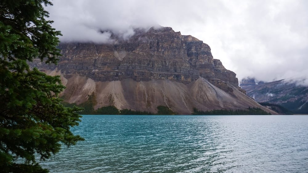 A wide view of Bow Lake's turquoise water in Banff National Park, Alberta, with a rugged, cloud-wrapped mountain rising above the shoreline along the Icefields Parkway.