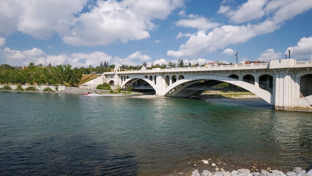 A view of the Bow River and a stone arch bridge from the riverside edge of Sien Lok Park in Calgary, Alberta, Canada, with a small boat passing beneath the arches and downtown buildings in the distance.