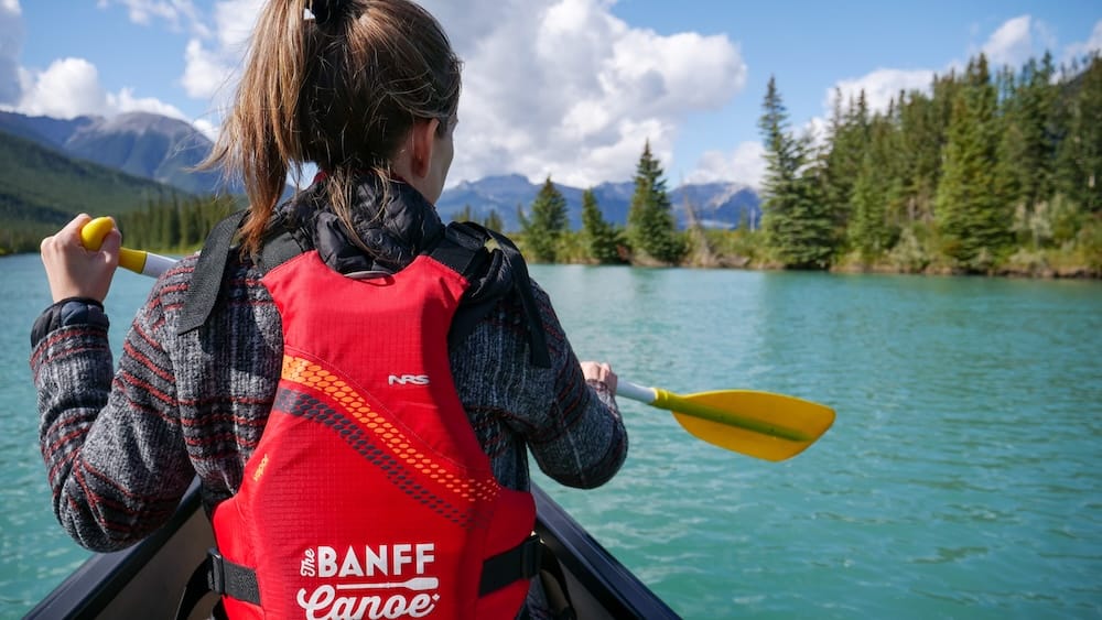 Audrey Bergner paddling the Bow River Big Canoe Tour in Banff, Alberta, Canada, with turquoise water and mountain views Audrey Bergner paddles on the Bow River during the Big Canoe Tour in Banff, Alberta, Canada, seen from behind in a red life jacket with a yellow paddle, turquoise water, evergreen forest and distant Rocky Mountain peaks ahead.