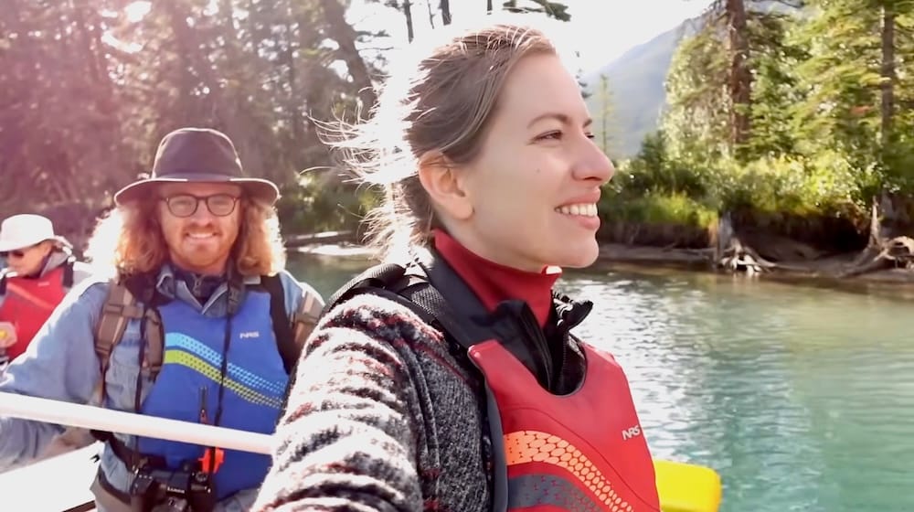 Audrey Bergner y Samuel Jeffery en el Big Canoe Tour por el río Bow en Banff, Alberta, Canadá Audrey Bergner sonriendo en primer plano con Samuel Jeffery detrás durante el Big Canoe Tour por el río Bow en Banff, Alberta, Canadá, ambos con chalecos salvavidas y rodeados de agua y bosque.