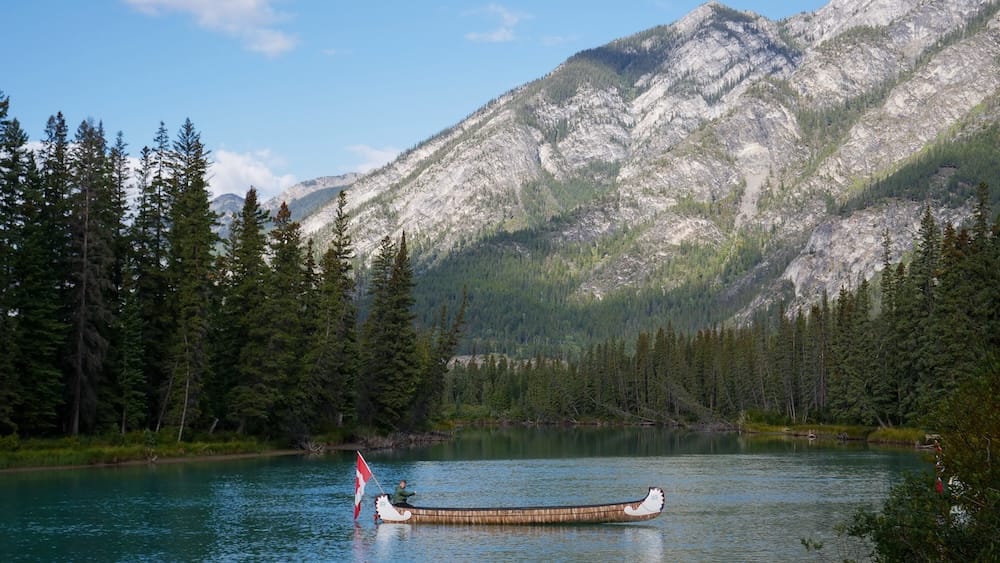 Big canoe on the Bow River in Banff, Alberta, Canada, during a Banff Canoe Club guided paddling tour A large Banff Canoe Club canoe glides along the turquoise Bow River in Banff, Alberta, Canada, with a Canadian flag at the stern and evergreen forest and gray Rocky Mountain slopes rising behind it.