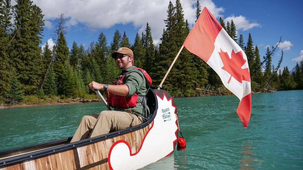 Guía del Big Canoe Tour en el río Bow, Banff, Alberta, Canadá, con canoa del Banff Canoe Club y bandera canadiense Un guía del Banff Canoe Club remando el Big Canoe sobre las aguas turquesas del río Bow en Banff, Alberta, Canadá, con una gran bandera canadiense, bosque de coníferas y cielo azul detrás.