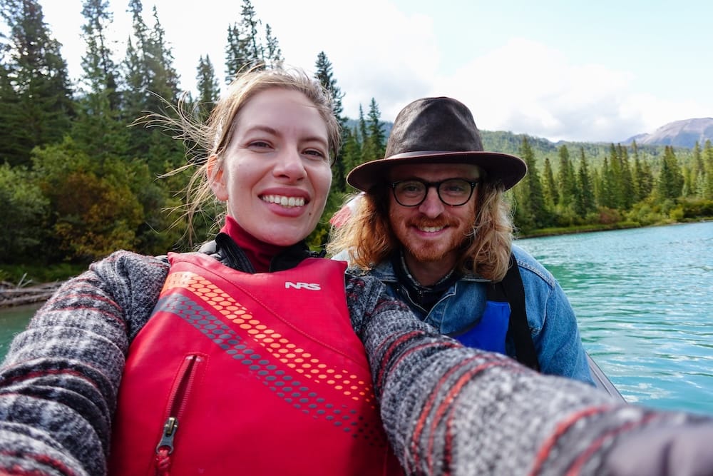 Samuel Jeffery y Audrey Bergner sonriendo con chalecos salvavidas a bordo del Big Canoe Tour guiado en el agua turquesa del río Bow cerca de Banff, Alberta, Canadá, con orillas boscosas y montañas detrás de ellos.