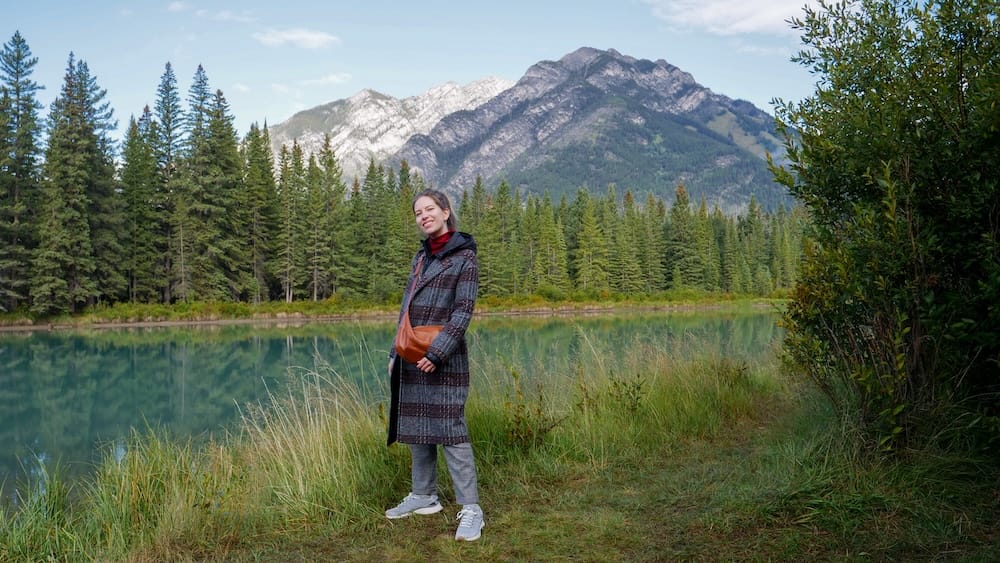 Audrey Bergner stands beside the Bow River in Banff, Alberta, Canada, with calm water, evergreen forest and layered Rocky Mountain peaks rising behind the riverside path.