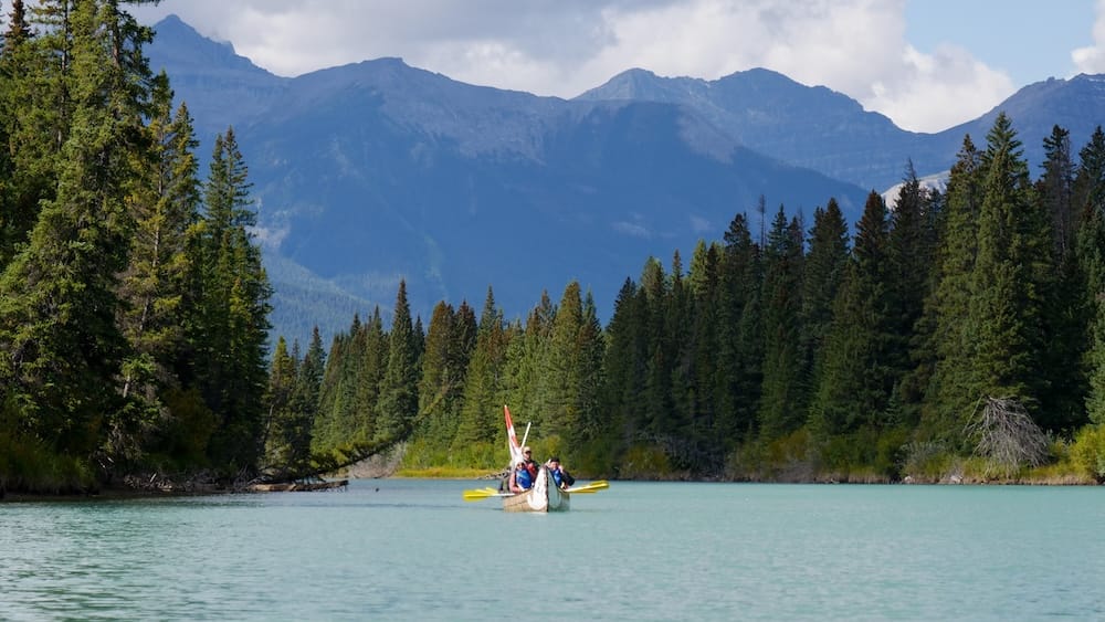 Grupo en Big Canoe guiado sobre el río Bow en Banff, Alberta, Canadá, con bosque y montañas de fondo Un grupo en Big Canoe guiado aparece a la distancia sobre el río Bow en Banff, Alberta, Canadá, con agua turquesa en primer plano, bosque de coníferas y laderas de las Montañas Rocosas detrás.