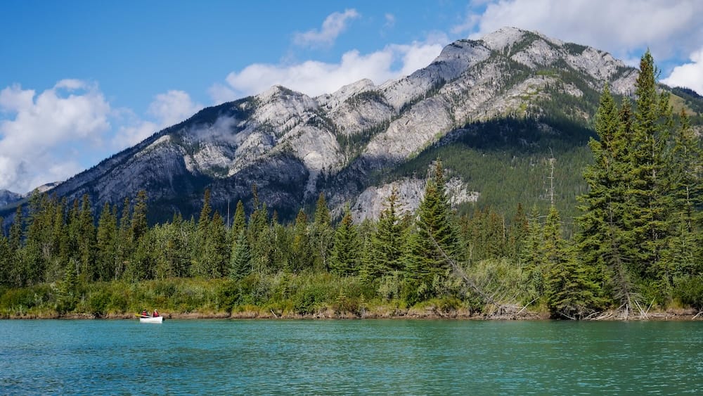 Independent canoeing on the Bow River in Banff, Alberta, Canada, with turquoise water and mountain scenery A small canoe paddles across the turquoise Bow River in Banff, Alberta, Canada, beneath forested banks and rugged Rocky Mountain slopes, showing an independent canoeing scene rather than a guided group format.
