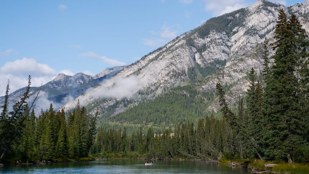 Una canoa a lo lejos en el tranquilo río Bow cerca de Banff, Alberta, Canadá, rodeada por un denso bosque de pinos y una cordillera escarpada con manchas de nieve bajo un cielo parcialmente nublado.