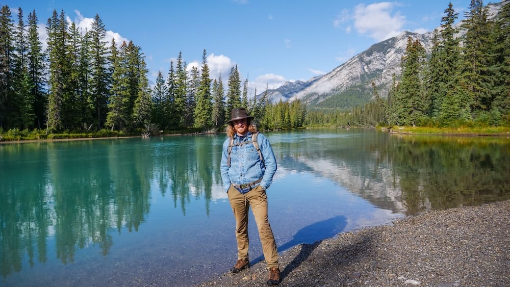 Bow River in Banff, Alberta, Canada with Samuel Jeffery beside the water on a relaxed first-day stop Samuel Jeffery stands on the rocky shore of the Bow River in Banff, Alberta, Canada, with turquoise water, conifer forest and mountain slopes behind him, illustrating the lower-pressure town-and-river start that works well on a three-day first visit.