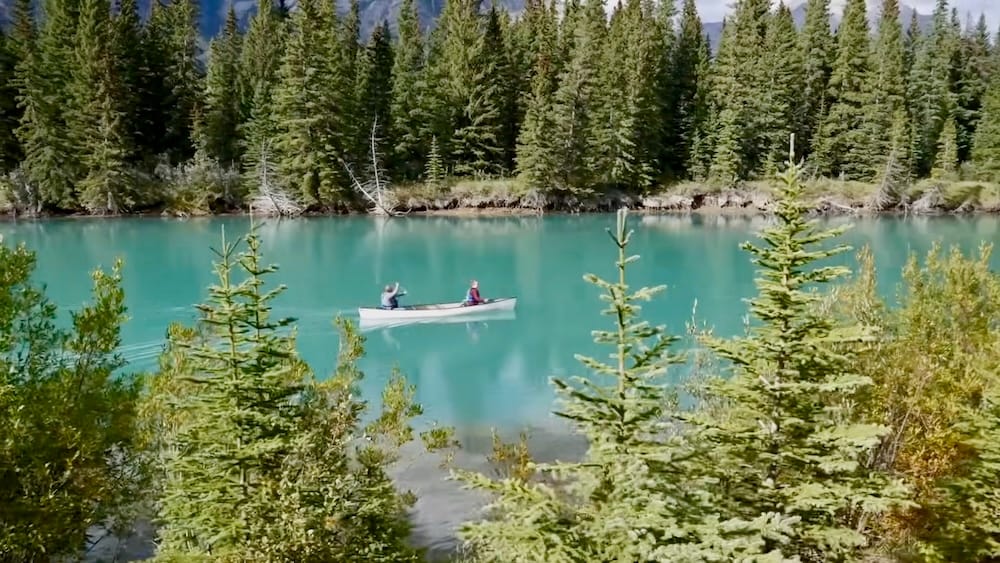 Vista de una canoa en el río Bow desde el paseo en carreta de Banff, Parque Nacional Banff, Alberta, Canadá Una canoa con dos personas cruza las aguas turquesas del río Bow en Banff, Alberta, Canadá, enmarcada por bosque de coníferas y vista desde la ruta del paseo en carreta del Cowboy Cookout.