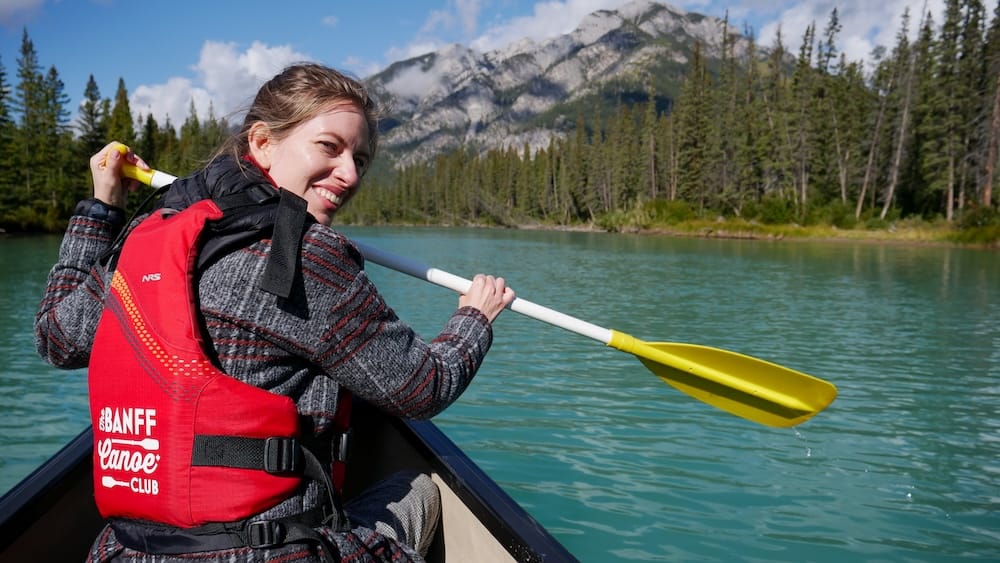 Audrey Bergner Paddling on the Bow River With Banff Canoe Club in Banff National Park, Alberta, Canada Audrey Bergner paddling a Big Canoe on the Bow River in Banff National Park, Alberta, Canada, wearing a Banff Canoe Club life jacket with forested mountains rising behind her.