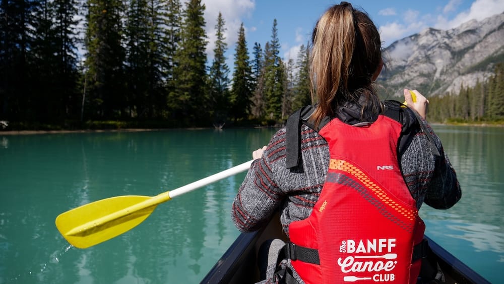 Bow River, Banff National Park, Alberta, Canada - Audrey Bergner paddling during a guided Big Canoe experience Audrey Bergner paddles a canoe on the Bow River in Banff National Park, Alberta, Canada, wearing a red Banff Canoe Club life jacket with turquoise water, evergreen forest and rocky mountains ahead.