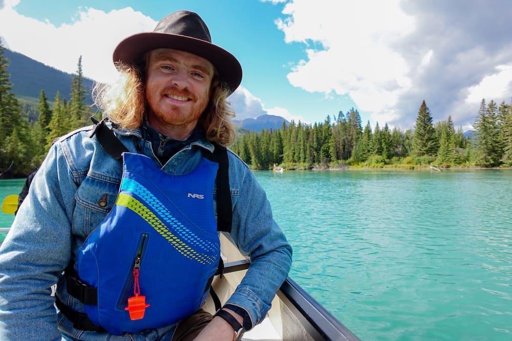 Bow River, Banff National Park, Alberta, Canada - Big Canoe tour with Samuel Jeffery on turquoise water Samuel Jeffery sits in a Big Canoe on the Bow River in Banff National Park, Alberta, Canada, wearing a blue life jacket and dark hat, with turquoise water, evergreen forest and mountain slopes behind him.