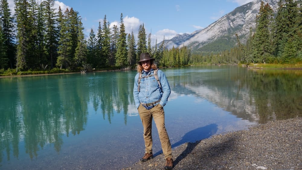 Samuel Jeffery junto al Bow River en el Parque Nacional Banff, Alberta, Canadá, con paisaje de las Montañas Rocosas Samuel Jeffery junto al Bow River de aguas turquesas, en el Parque Nacional Banff, Alberta, Canadá, con bosque de coníferas, agua calma y picos de las Montañas Rocosas detrás.