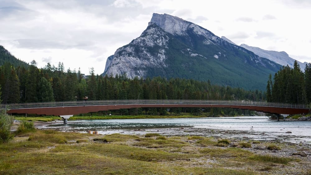 Bow River flowing through Banff, Alberta, Canada, beneath a pedestrian bridge with forested banks and a steep Rocky Mountain rising behind it under an overcast sky.