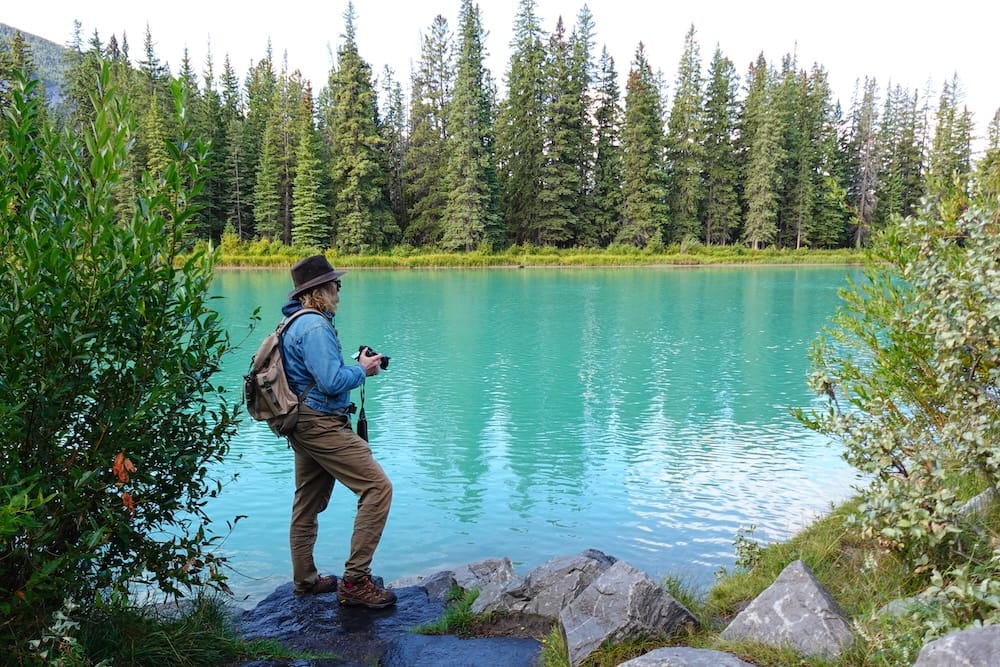 Samuel Jeffery photographing the turquoise Bow River in Banff town, Alberta, Canada, with evergreen forest lining the opposite bank during a free riverside walk.