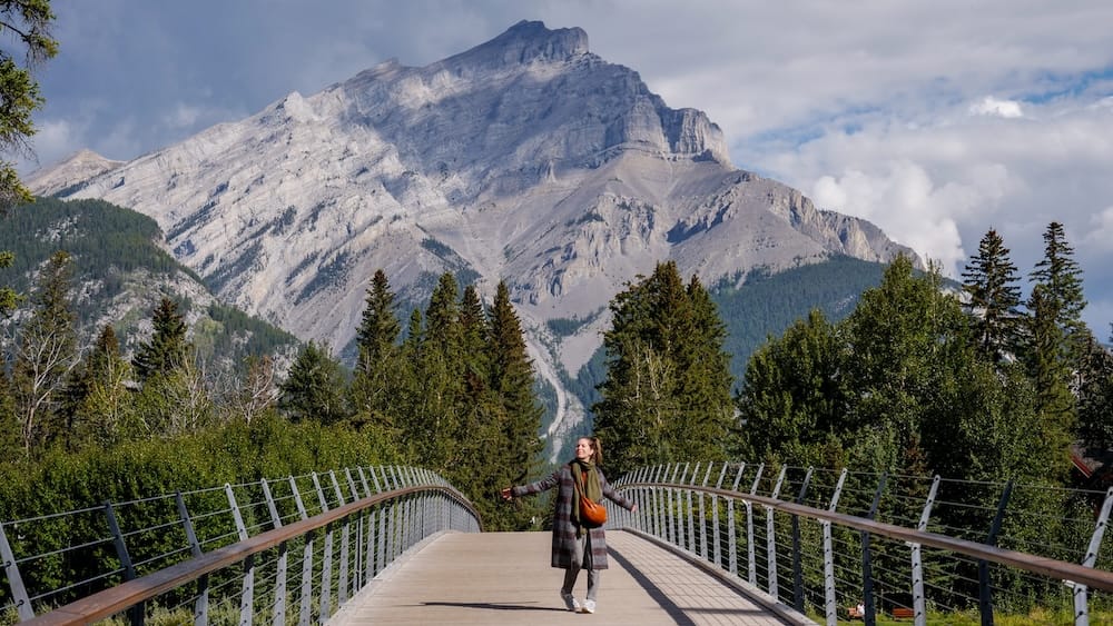 Audrey Bergner on a Bow River Bridge near Central Park in Banff, Alberta, Canada, with Mountain Views Audrey Bergner standing on a Bow River pedestrian bridge near Central Park in Banff, Alberta, Canada, with dense trees and a massive rocky Canadian Rockies peak filling the background.