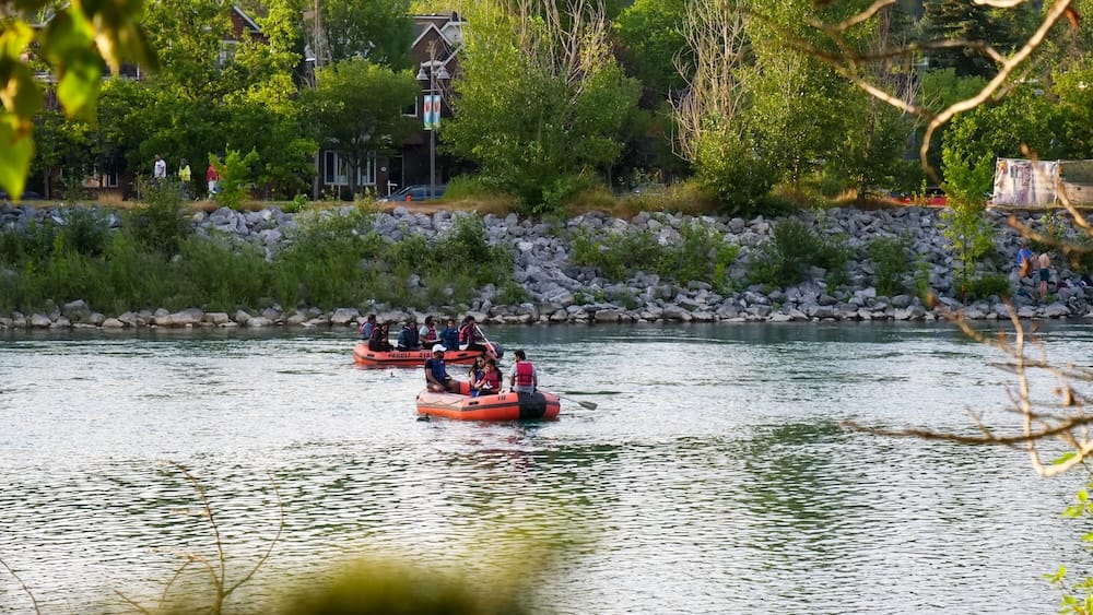 Bow River Rafting in Calgary Alberta Canada with Summer Paddlers Along the Urban Riverfront Two inflatable rafts carrying paddlers move along the Bow River in Calgary, Alberta, Canada, with a rocky shoreline, dense summer greenery and riverside buildings in the background.