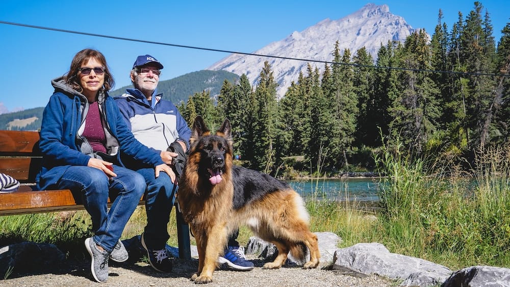 Riverside bench stop on the Bow River Trail in Banff, Alberta, Canada, with Togo the German Shepherd and mountain views Daniel and Maria Bergner sit on a riverside bench along the Bow River Trail in Banff, Alberta, Canada, with their German Shepherd Togo beside them and a snow-capped Rocky Mountain peak rising behind the forest and river.