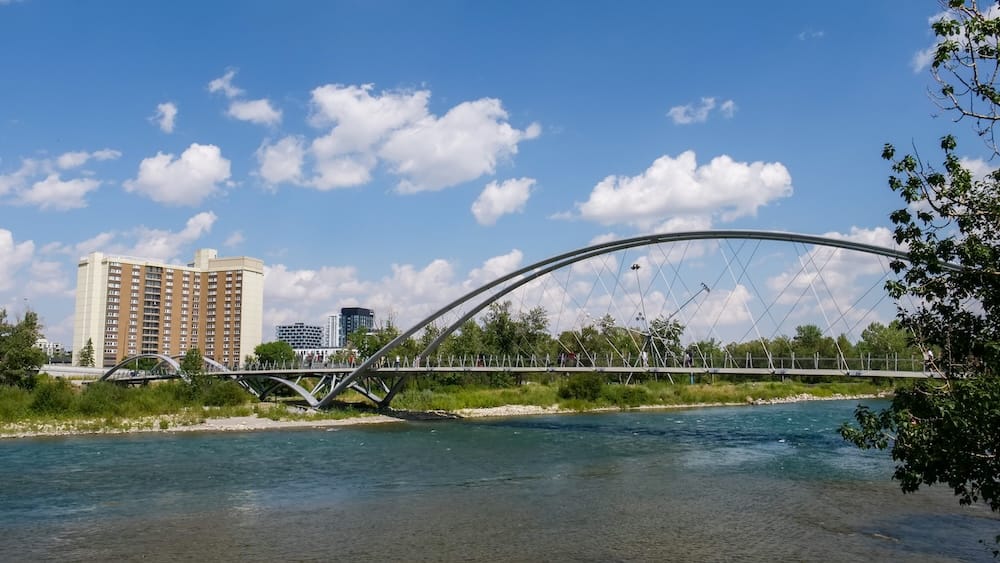 Vista de verano sobre el Bow River en Calgary, Alberta, Canadá, con un gran puente peatonal arqueado sobre el agua, riberas verdes y edificios de la ciudad bajo un cielo azul despejado.