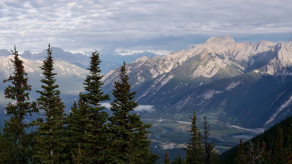 Layered evergreen trees frame a wide view of the Bow Valley and surrounding Rocky Mountain ranges under a partly cloudy sky, seen from Sulphur Mountain near Banff, Alberta, Canada.