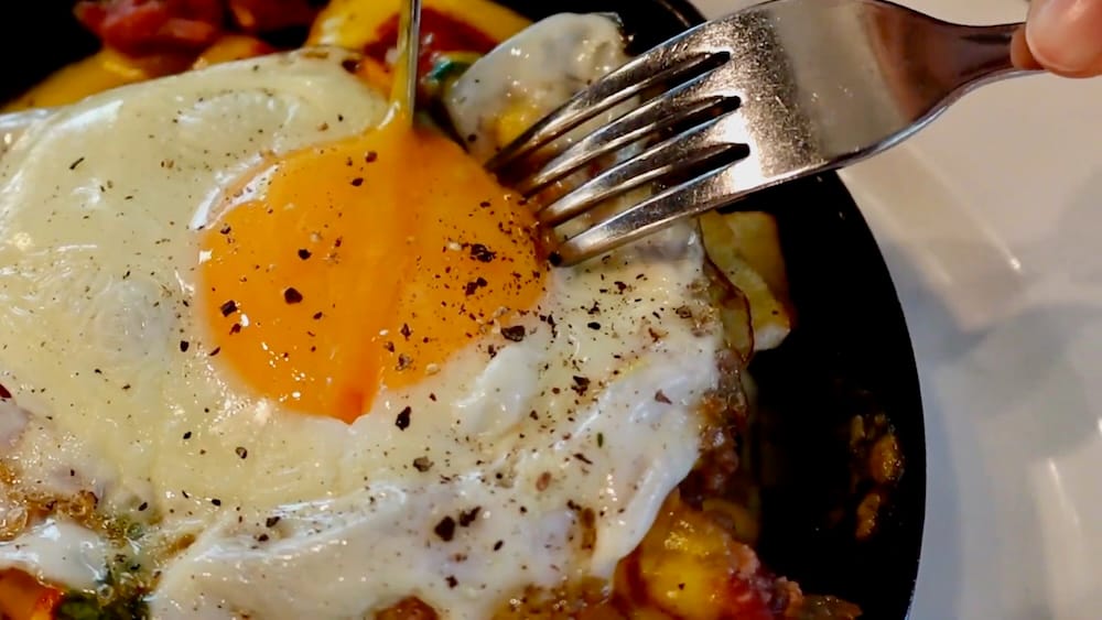 Runny Egg Yolk on Breakfast Poutine at The Beltliner, Beltline, Calgary, Alberta, Canada Close-up of a fork breaking a runny sunny-side-up egg yolk over hash browns and cheese in a cast-iron skillet at The Beltliner in Beltline, Calgary, Alberta, Canada.