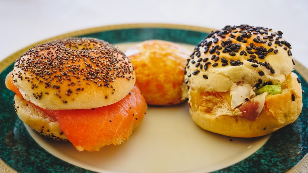 Brie, Jamón Crudo, and Avocado Bagel at Llao Llao Hotel Afternoon Tea, Bariloche A plate with a salmon poppy seed bagel, a small cheese scone, and a black sesame bagel filled with brie, jamón crudo, and avocado, part of the savory course during afternoon tea at Llao Llao Hotel in Bariloche, Río Negro, Argentine Patagonia.