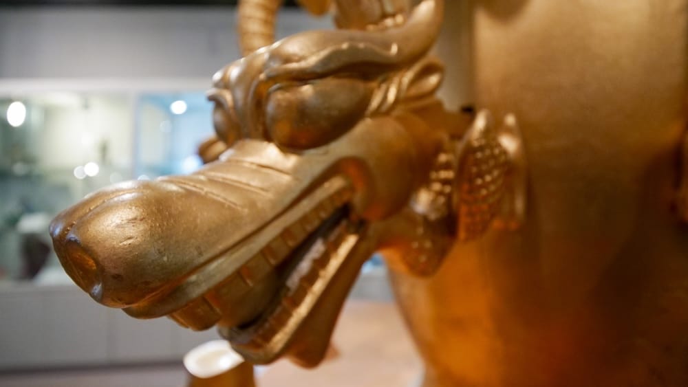 Close-up macro detail of a gilded bronze dragon head, part of a larger bronze artifact on display in the Chinese Artifacts Museum in Calgary, Alberta, Canada, with museum display cases visible softly in the background.