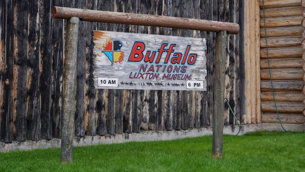Wooden Buffalo Nations Luxton Museum sign outside the log-built museum in Banff, Alberta, Canada, showing the institution’s former name against rustic timber walls and green grass.