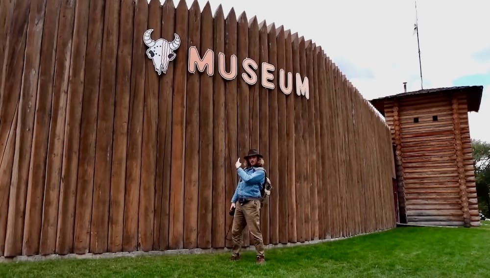 Samuel Jeffery afuera de Buffalo Nations Museum en Banff, Alberta, Canadá, junto a la empalizada de madera Samuel Jeffery parado afuera de Buffalo Nations Museum en Banff, Alberta, Canadá, junto a la alta empalizada de madera y el cartel del museo, con una torre de troncos al fondo.