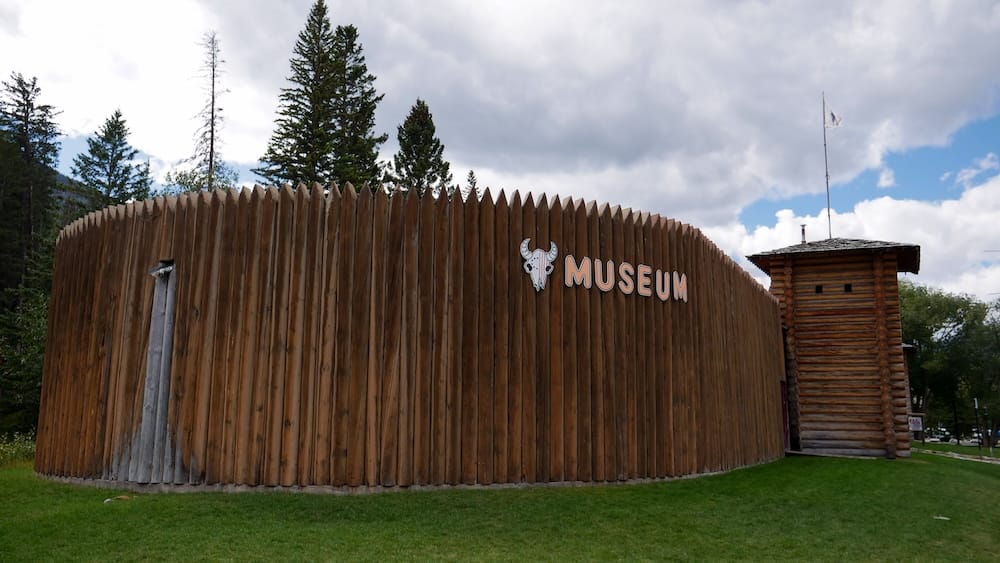 Exterior de Buffalo Nations Museum con empalizada y torre de troncos en Banff, Alberta, Canadá Vista amplia del exterior de Buffalo Nations Museum en Banff, Alberta, Canadá, con la alta empalizada de madera, el cartel del museo, una torre de troncos, césped y cielo nublado.