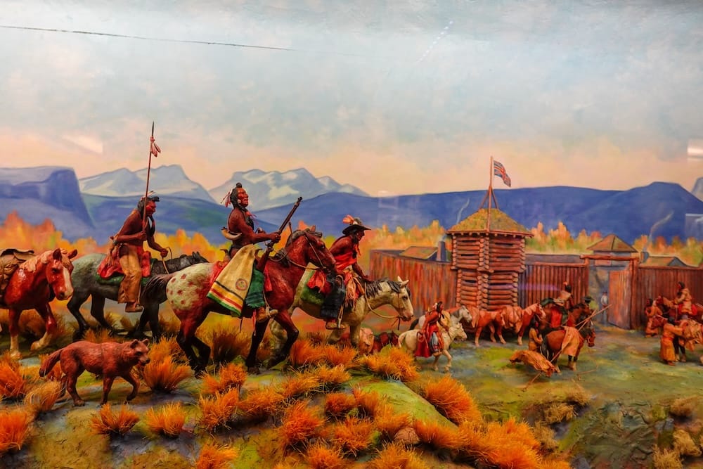 Diorama histórico con jinetes indígenas y fuerte en Buffalo Nations Museum, Banff, Alberta, Canadá Diorama histórico detallado dentro de Buffalo Nations Museum en Banff, Alberta, Canadá, con jinetes indígenas acercándose a un fuerte de madera frente a un fondo pintado de montañas.