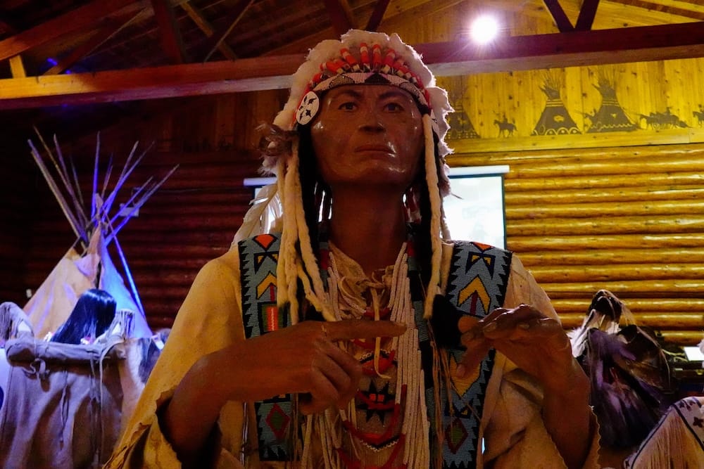Figura indígena con trabajos de cuentas y tocado de plumas en Buffalo Nations Museum, Banff, Alberta, Canadá Figura indígena detallada con trabajos de cuentas y tocado de plumas dentro de Buffalo Nations Museum en Banff, Alberta, Canadá, con un tipi y otras exhibiciones culturales detrás.
