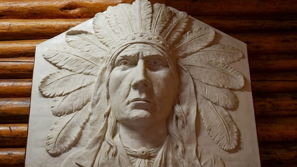 Figura indígena esculpida con tocado de plumas dentro de Buffalo Nations Museum en Banff, Alberta, Canadá Figura indígena esculpida con un gran tocado de plumas expuesta contra paredes de troncos dentro de Buffalo Nations Museum en Banff, Alberta, Canadá.