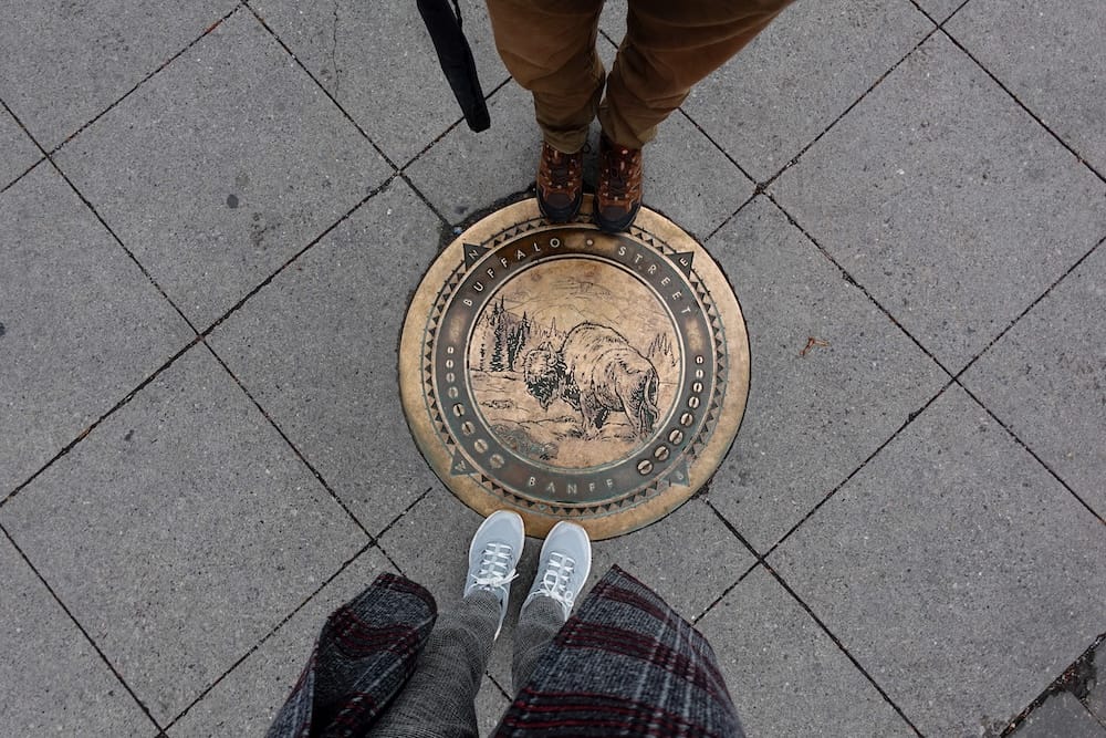 Buffalo Street marker in Banff town, Alberta, Canada, with Samuel Jeffery and Audrey Bergner Samuel Jeffery and Audrey Bergner stand over a decorative Buffalo Street marker embedded in the pavement in Banff town, Alberta, Canada, with only their shoes and lower legs visible.
