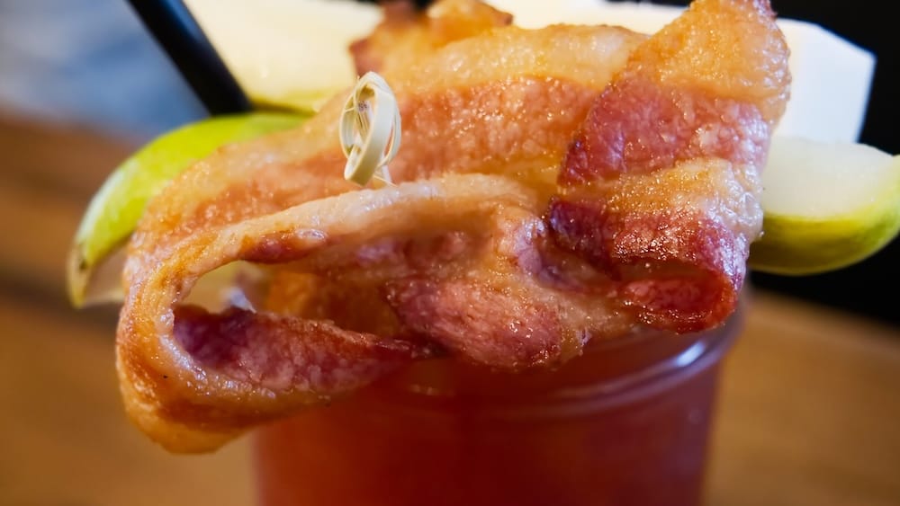 Caesar Cocktail with Bacon Garnish at The Beltliner, a Diner in the Beltline, Calgary, Alberta, Canada Close-up of a Caesar cocktail at The Beltliner in Calgary topped with a thick woven bacon garnish and lime wedges, the Clamato-red drink visible beneath.