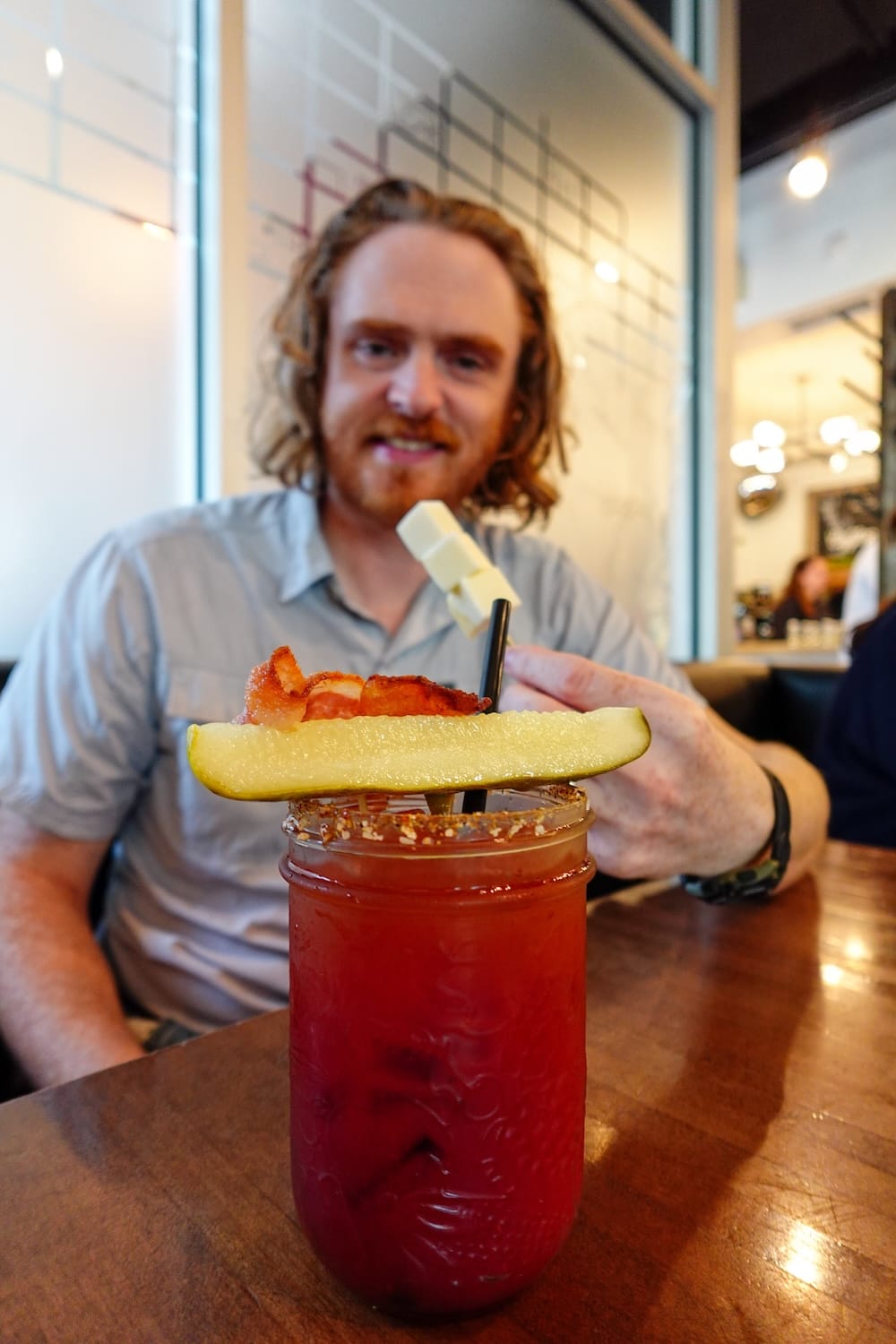 Samuel Jeffery sentado detrás de un Caesar rojo en Calgary, Alberta, Canadá, servido en un frasco con borde condimentado y una gran guarnición de pepinillo, panceta y queso.