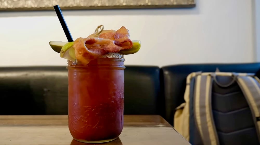 Un Caesar en Calgary, Alberta, Canadá, servido en un frasco de vidrio sobre una mesa de restaurante, con guarnición de panceta y pepinillo, borde condimentado y una pajita negra sobre la bebida roja.