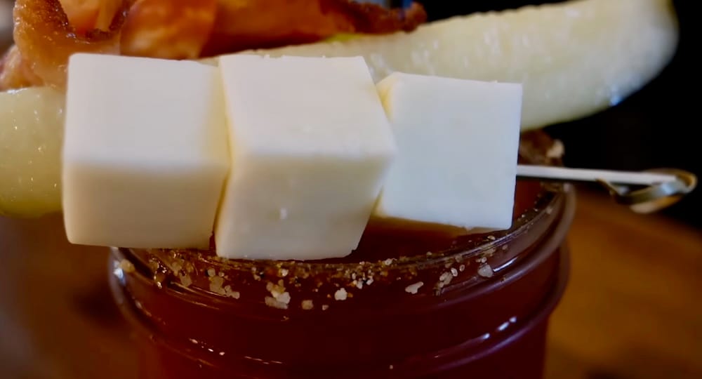 Primer plano de un Caesar en Calgary, Alberta, Canadá, con cubos de queso, un pepinillo, panceta y un borde con condimentos gruesos sobre la bebida roja.