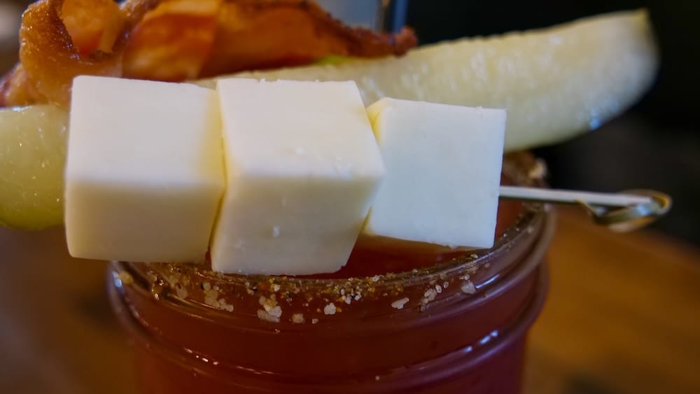 Macro close-up of a Caesar cocktail in Calgary, Alberta, Canada, showing the red drink, seasoned rim and oversized garnish with cheese cubes, pickle and bacon arranged across the top.