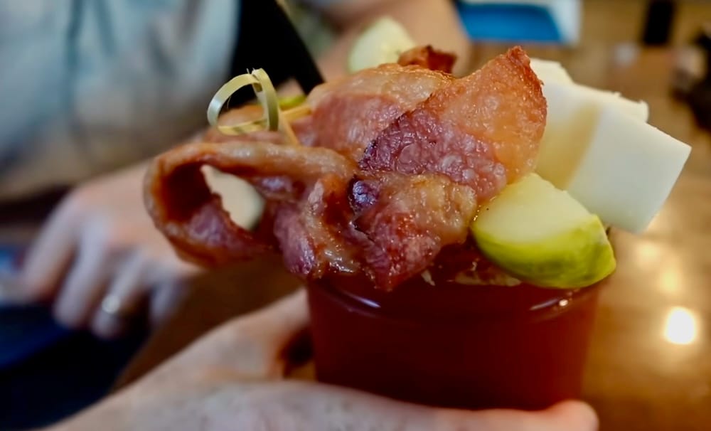 Samuel Jeffery sostiene un Caesar en Calgary, Alberta, Canadá, con la bebida roja de brunch llenando el primer plano y una guarnición de panceta, queso y pepinillo apilada sobre el borde.