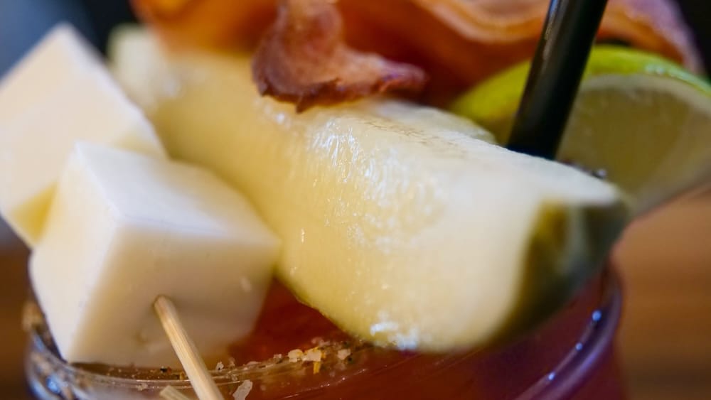 Primerísimo plano de un Caesar en Calgary, Alberta, Canadá, con un pepinillo grande, cubos de queso blanco, lima, panceta y parte del borde condimentado sobre la bebida roja y salada.