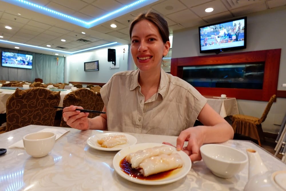 Audrey Bergner sits at a restaurant table eating dim sum in Calgary, Alberta, Canada, with plates of soft rice rolls, chopsticks and tea visible in front of her during one of the city’s standout Chinese food meals.