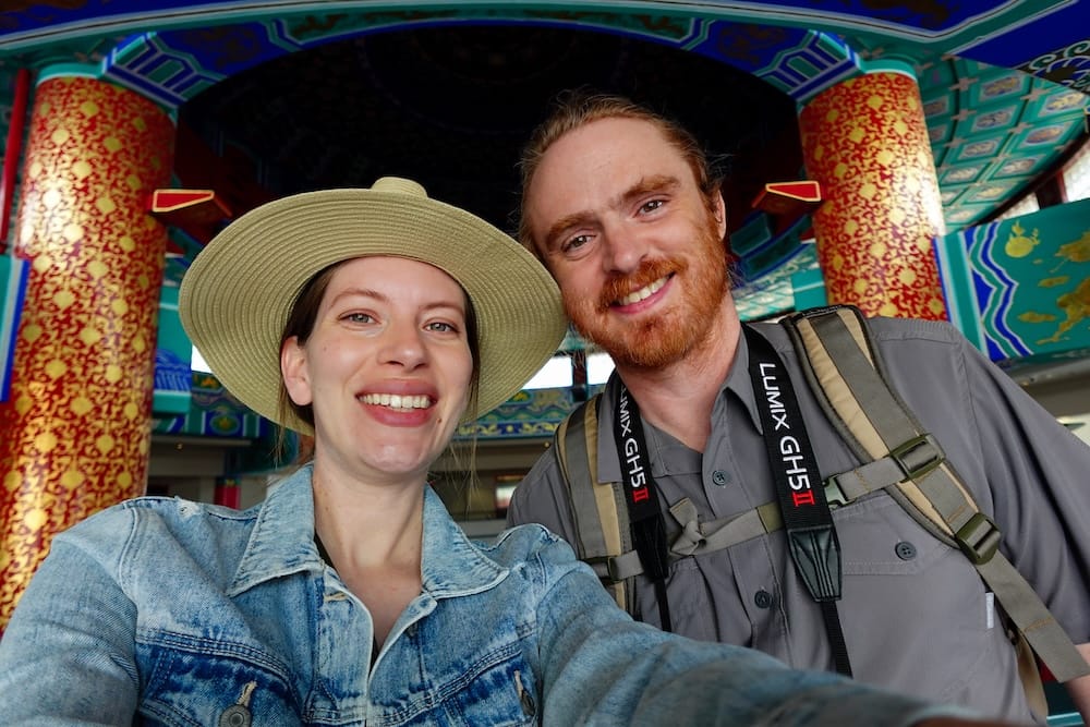 Samuel Jeffery y Audrey Bergner sonriendo juntos durante su viaje por Calgary, Alberta, Canadá, con detalles arquitectónicos coloridos detrás y Samuel llevando equipo fotográfico.