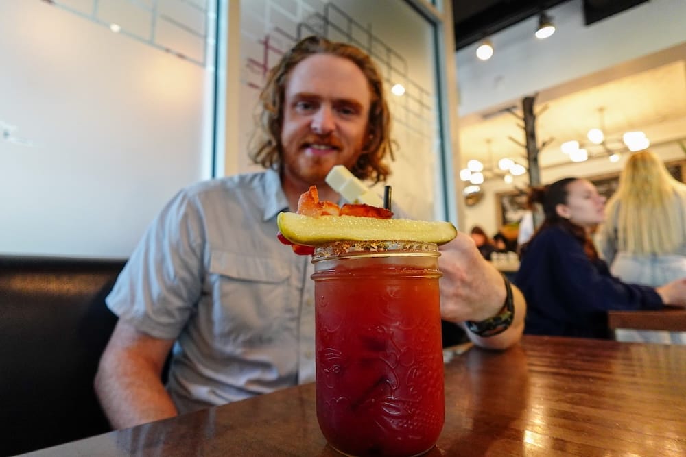 Samuel Jeffery sits at a restaurant table in Calgary, Alberta, Canada, with an elaborate Caesar cocktail in a mason jar garnished with a pickle spear, bacon, and salt rim in the foreground.