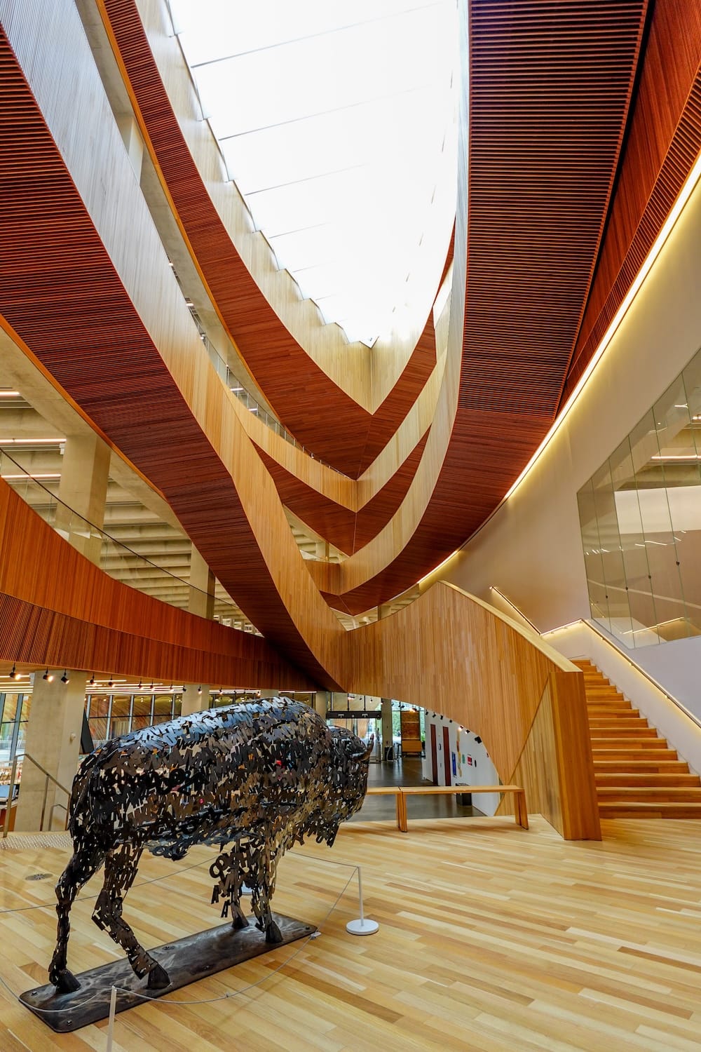 Atrio de madera y escultura de bisonte en Calgary Central Library, Calgary, Alberta, Canadá Interior de Calgary Central Library en Calgary, Alberta, Canadá, con balcones curvos de madera, un atrio iluminado desde arriba, escaleras de madera y una gran escultura metálica de un bisonte en primer plano.