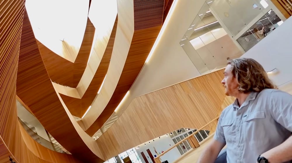 Samuel Jeffery admirando la arquitectura de madera dentro de Calgary Central Library, Calgary, Alberta, Canadá Samuel Jeffery mira hacia arriba la arquitectura curva de madera dentro de Calgary Central Library, Calgary, Alberta, Canadá, con balcones de madera, barandas de vidrio y un interior luminoso sobre él.