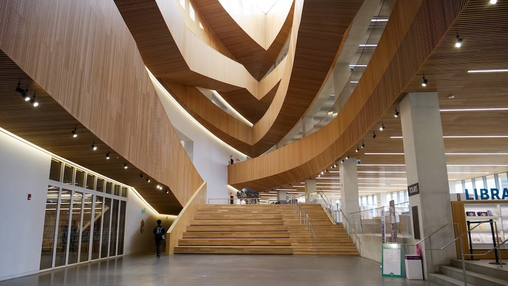 Interior of Calgary Central Library in downtown Calgary, Alberta, Canada, showing the sweeping wood-lined ceiling, broad staircase, open public space, and upper levels of the library’s modern interior.