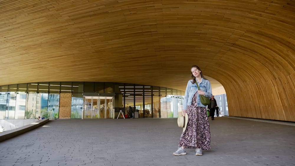Calgary Central Library East Village Alberta Canada Architecture with Audrey Bergner on Foot Audrey Bergner stands beneath the sweeping wooden entrance of Calgary Central Library in East Village, Calgary, Alberta, Canada, with glass doors and the curved timber ceiling behind her.