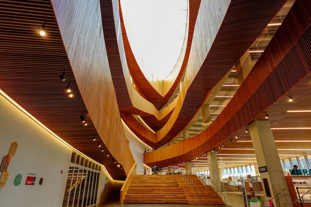 Calgary Central Library Interior in East Village Calgary Alberta Canada with Curved Wood Architecture and Atrium Interior of Calgary Central Library in East Village, Calgary, Alberta, Canada, showing sweeping wooden architectural lines, broad staircases, warm lighting and the open central atrium.