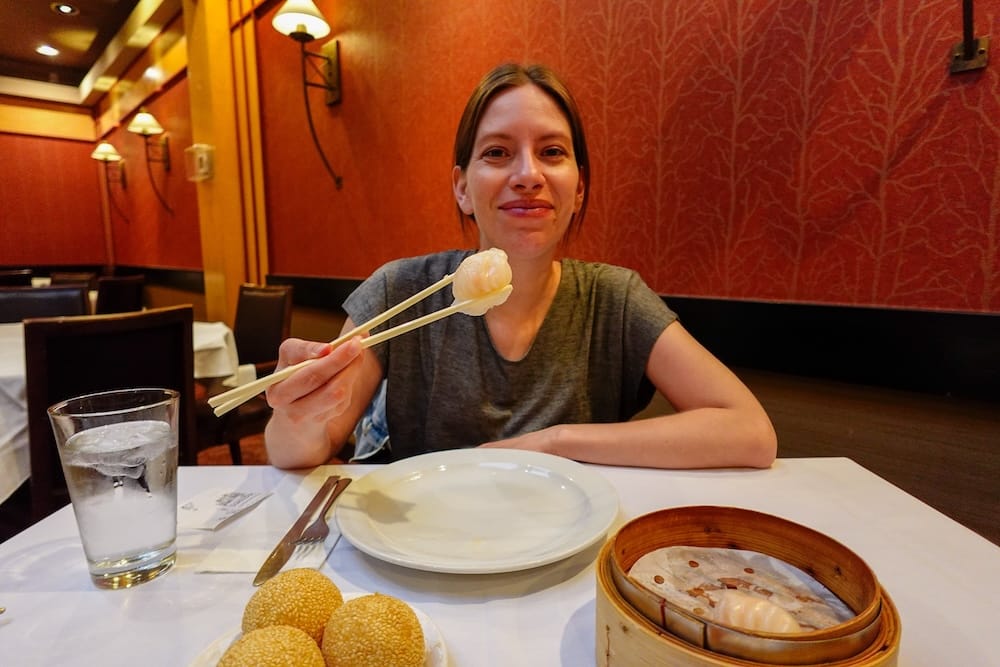 Audrey Bergner enjoying dim sum at a restaurant in Calgary Chinatown, Alberta, Canada, holding a dumpling with chopsticks at a table with a bamboo steamer basket and sesame balls.